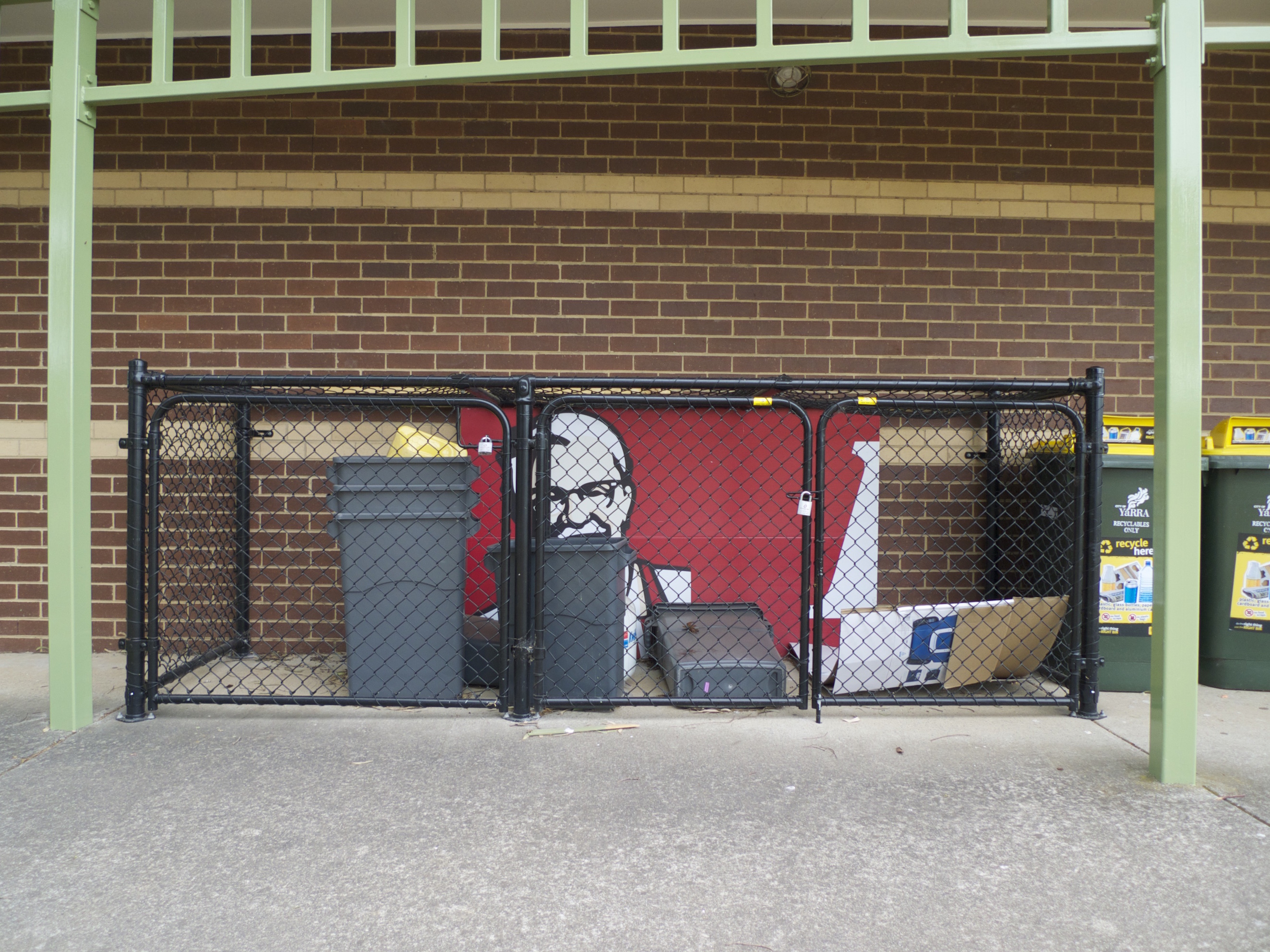 Un morceau d'une grande illustration KFC sous verrou dans un espace poubelle grillagé, devant un mur en briques