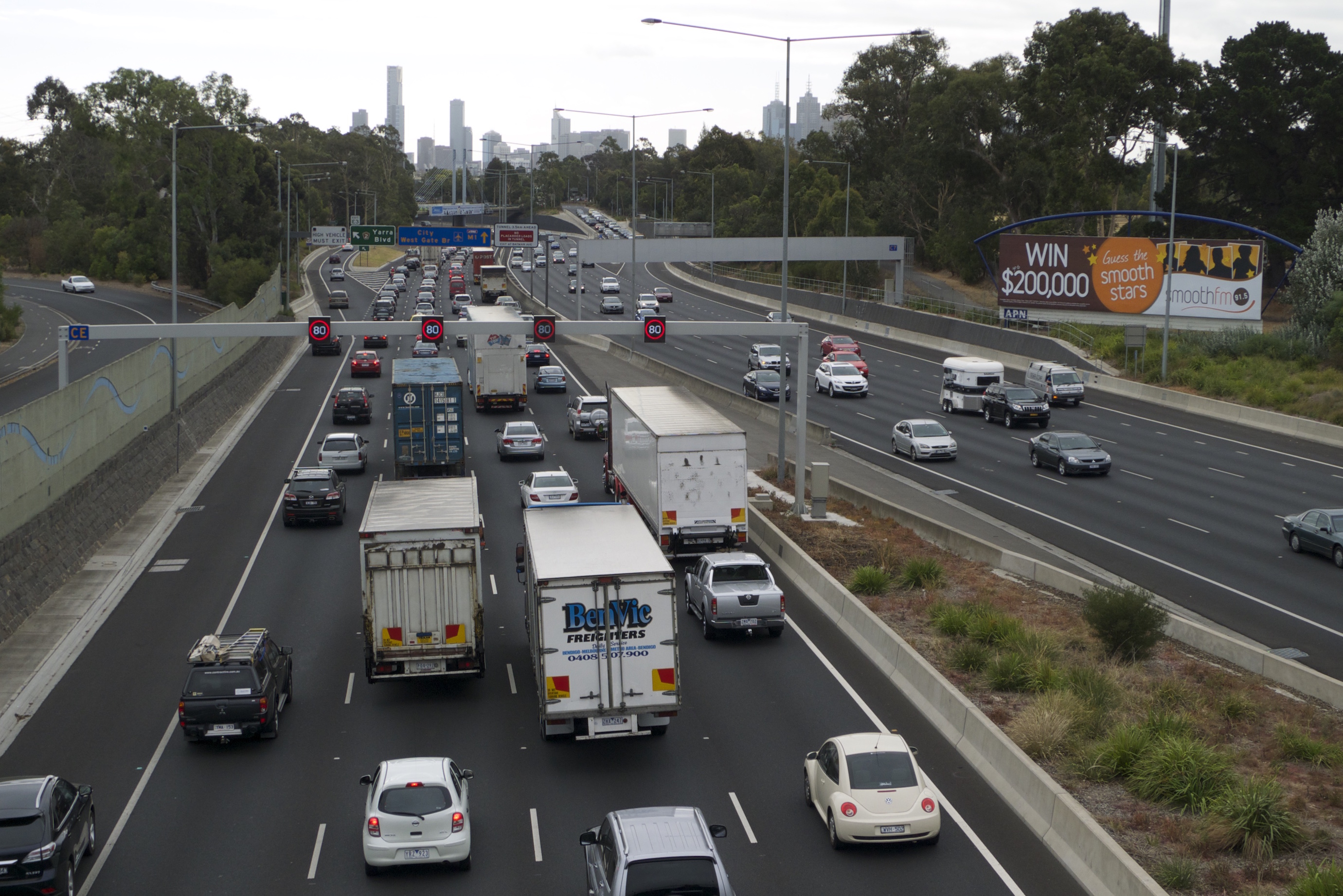 Autoroute à 4 voies dans les deux sens, conduite à gauche, vue depuis un pont, avec un embouteillage à gauche, vue sur les buildings du centre de Melbourne au loin