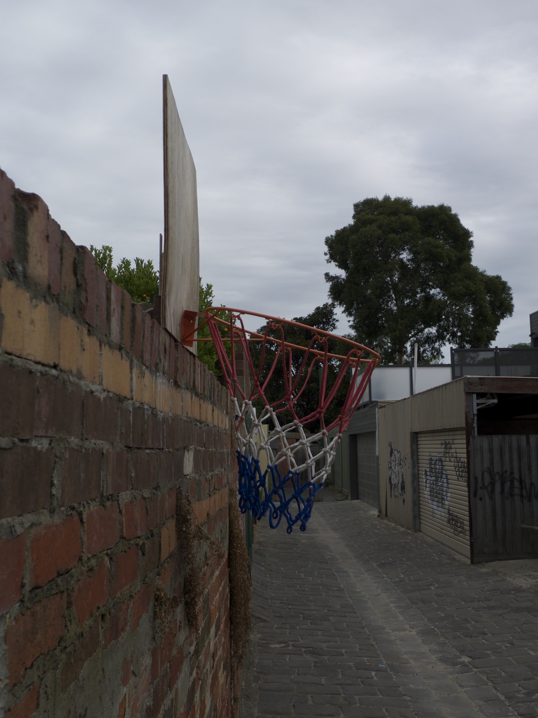 Panneau de basket vu de profil, au dessus d'un mur en briques dans une 'ruelle de derrière'
