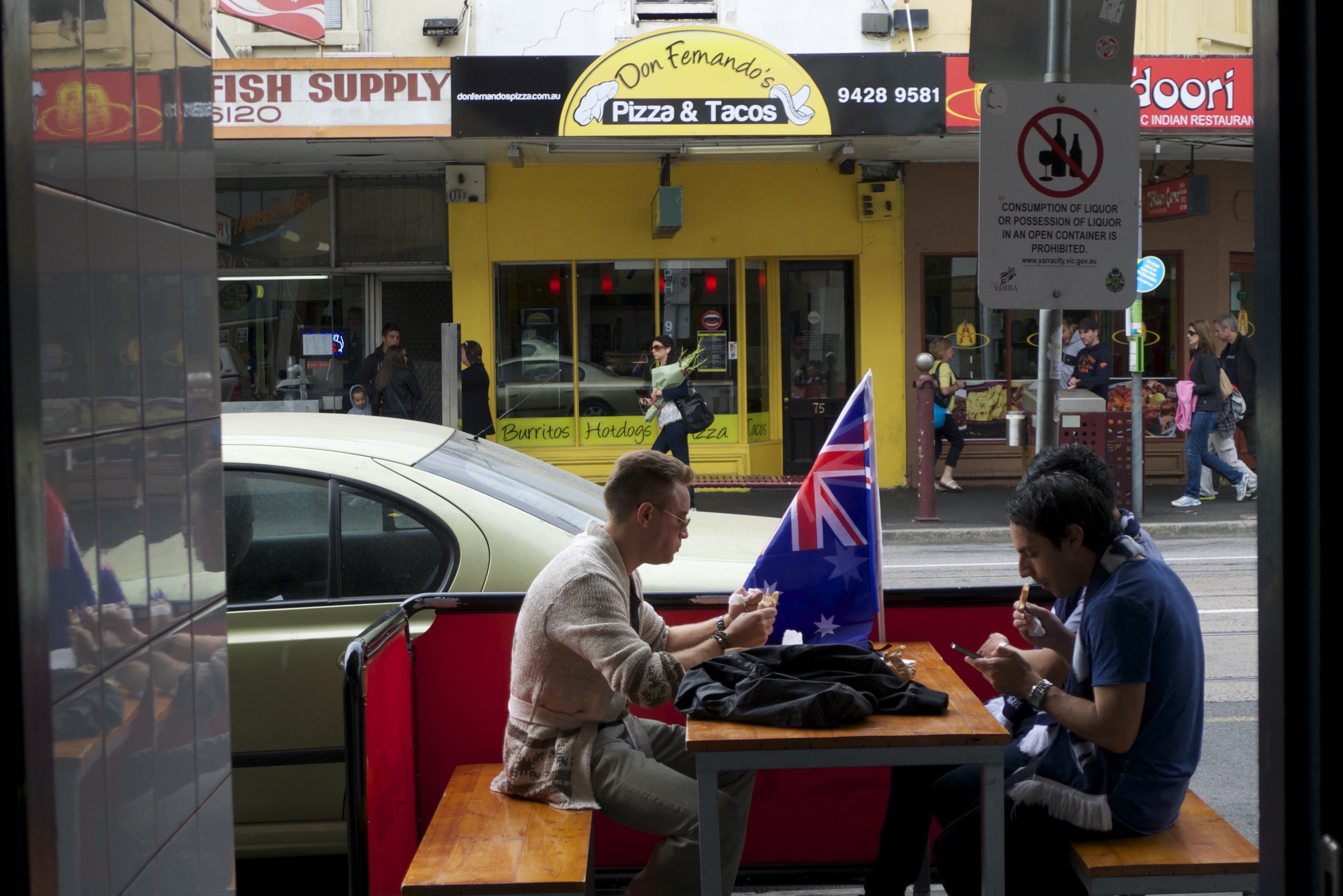 Trois personnes mangeant et regardant leurs téléphones installés à une table avec un drapeau australien
